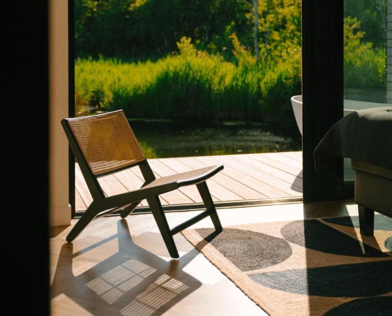 Zonnig interieur van een luxe vakantiehuis in Nederland met houten stoel, open schuifpui en uitzicht op het water en de natuur.
