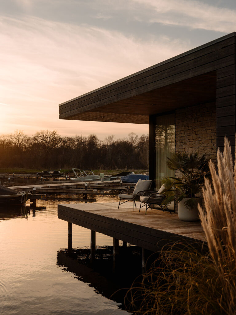 Die Terrasse einer modernen Wasser-Villa bei Sonnenuntergang, mit Sitzgelegenheiten am Wasser, warmem Licht und Blick auf einen ruhigen Yachthafen