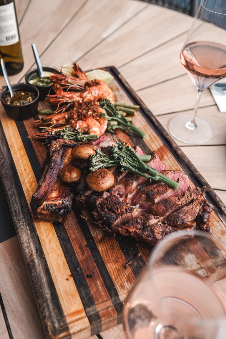 Eine großzügige Snackplatte mit gegrilltem Fleisch, Garnelen, Gemüse, Saucen und Rosé auf einem Tisch im Freien für ein stimmungsvolles kulinarisches Erlebnis am Wasser