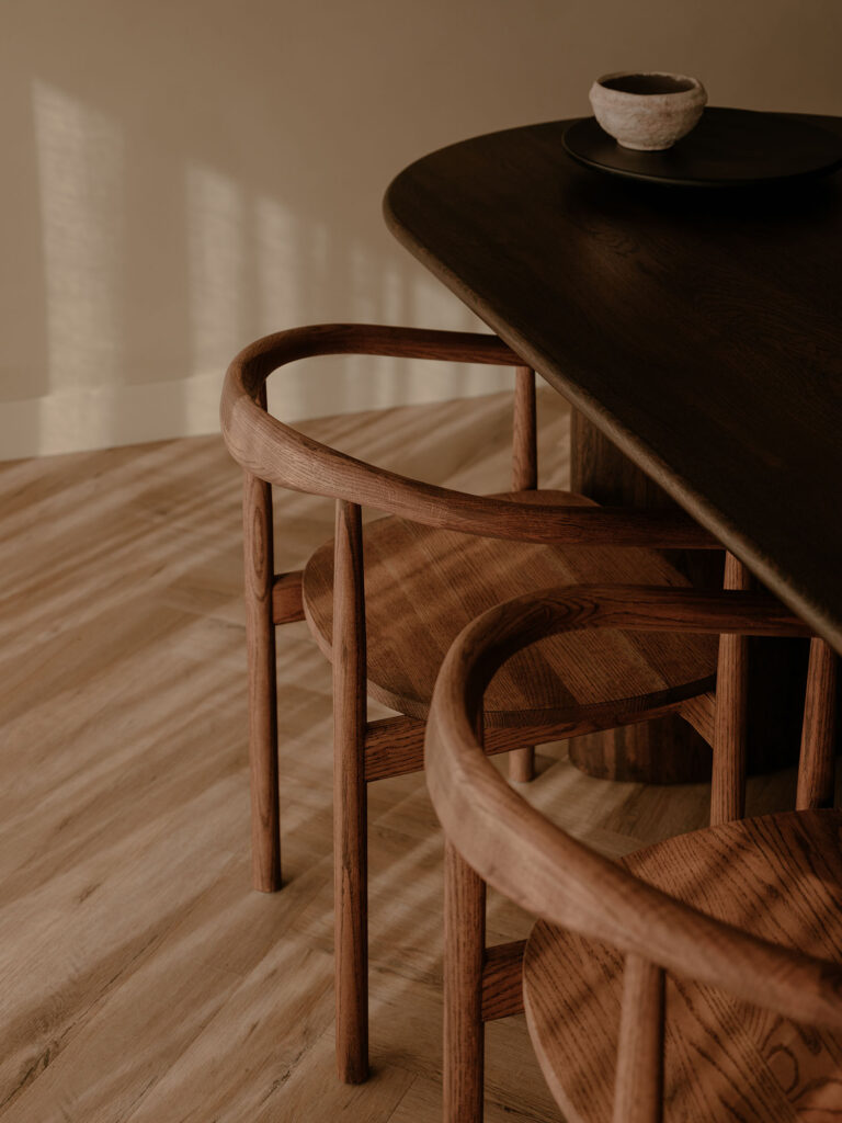 Ein Detail eines hölzernen Esstisches und Designerstühlen mit warmem Lichteinfall und natürlichen Materialien in einer ruhigen, stilvollen Umgebung