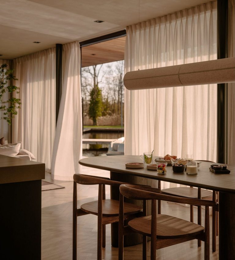 Heller Essbereich mit natürlichen Materialien, Holztisch und -stühlen, großen Fenstern und Vorhängen mit Blick auf das Wasser und eine ruhige, grüne Umgebung