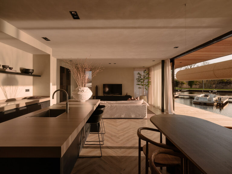 Ein stilvoller Wohnbereich mit offener Küche, Esstisch und sanften Naturtönen, der nahtlos in eine großzügige Terrasse am Wasser in einer ruhigen und nachhaltigen Umgebung übergeht