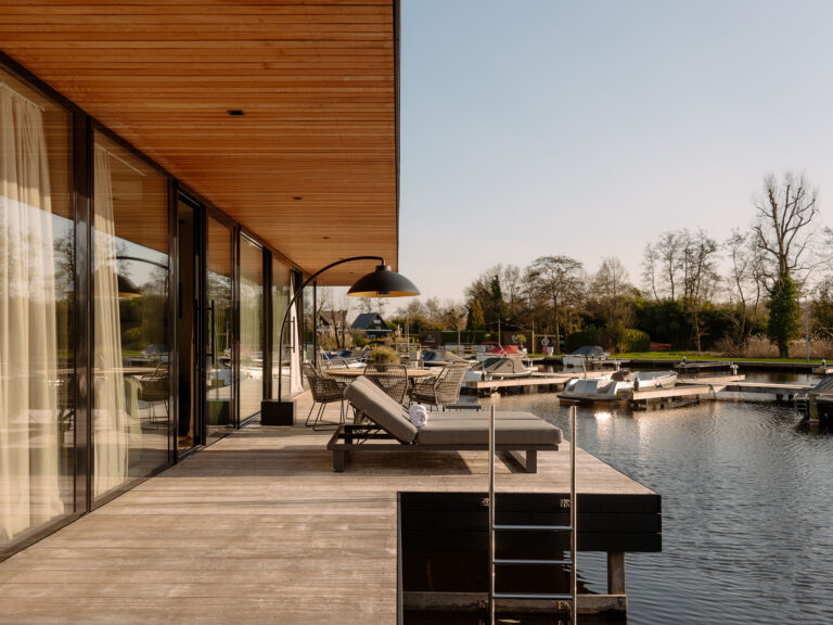 Ruim houten terras van een watervilla met ligbed, eettafel, zwemtrap en direct uitzicht op het water en de jachthaven