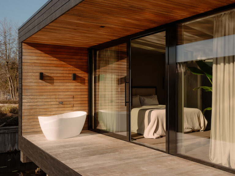 Sfeervolle slaapkamer met grote glazen schuifpuien naar een houten privé terras met vrijstaand buitenbad en warme houtafwerking aan het water