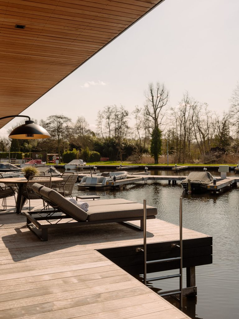 Stilvolle Terrasse einer Wasser-Villa mit Liegestuhl, Esstisch und direktem Zugang zum Wasser mit Anlegesteg sowie Blick auf einen ruhigen Yachthafen und eine naturreiche Umgebung