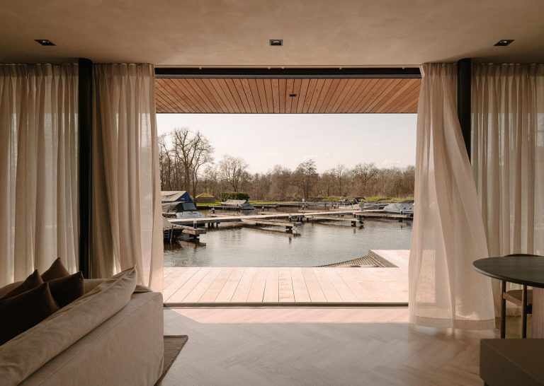 Helles Wohnzimmer mit Flügeltüren und Vorhängen, das auf eine Terrasse am Wasser mit Anlegestellen und Booten in einer ruhigen Yachthafenumgebung blickt