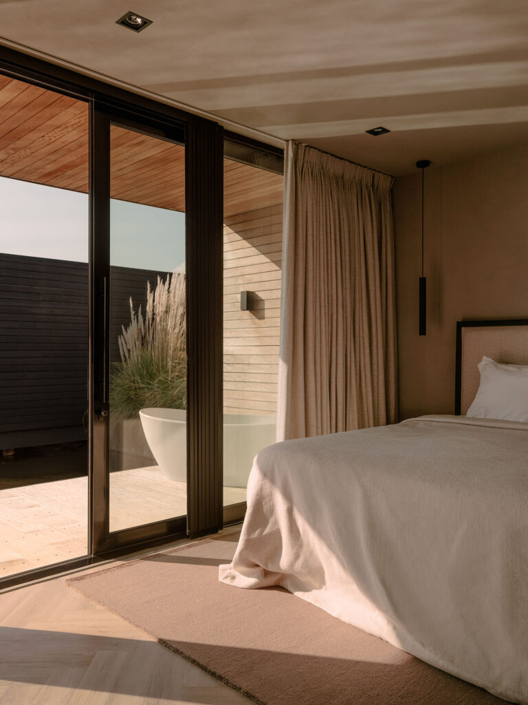 Ruhiges Schlafzimmer mit weicher Bettwäsche, warmen, neutralen Farbtönen und Flügeltüren, die auf eine private Terrasse mit Außenpool führen