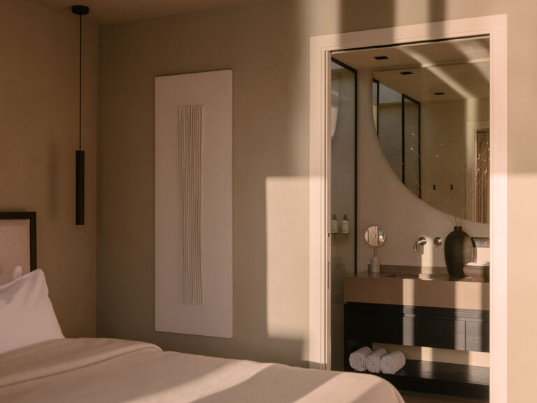 Sfeervolle slaapkamer met zachte neutrale tinten en doorkijk naar een stijlvolle badkamer met wastafel, ronde spiegel en warme lichtinval