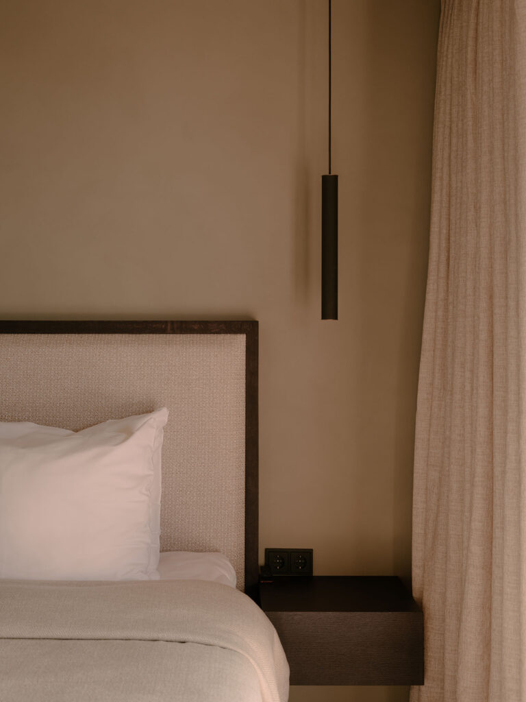 Detailansicht eines stilvollen Bettes mit gepolstertem Kopfteil, Leinenvorhängen und einer minimalistischen Hängelampe in warmen, neutralen Farbtönen, die eine ruhige und nachhaltige Atmosphäre schaffen