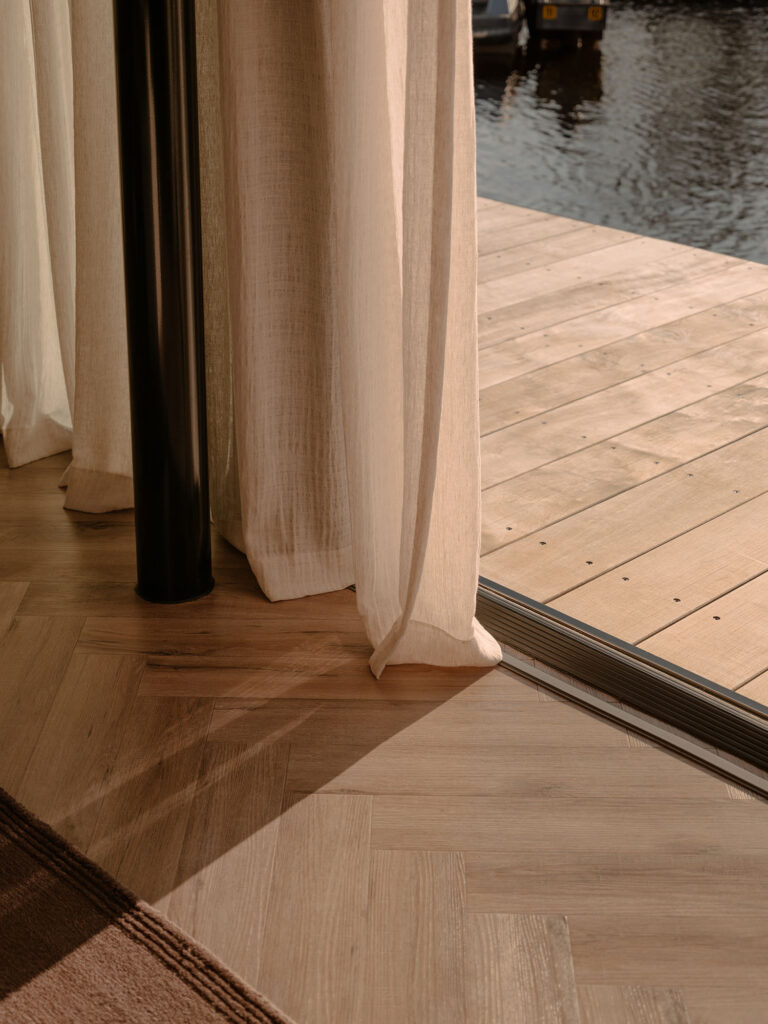 Ein Detail der Leinenvorhänge, des Fischgrätenparketts und des Übergangs zur Terrasse am Wasser in einer warmen, ruhigen Villenatmosphäre