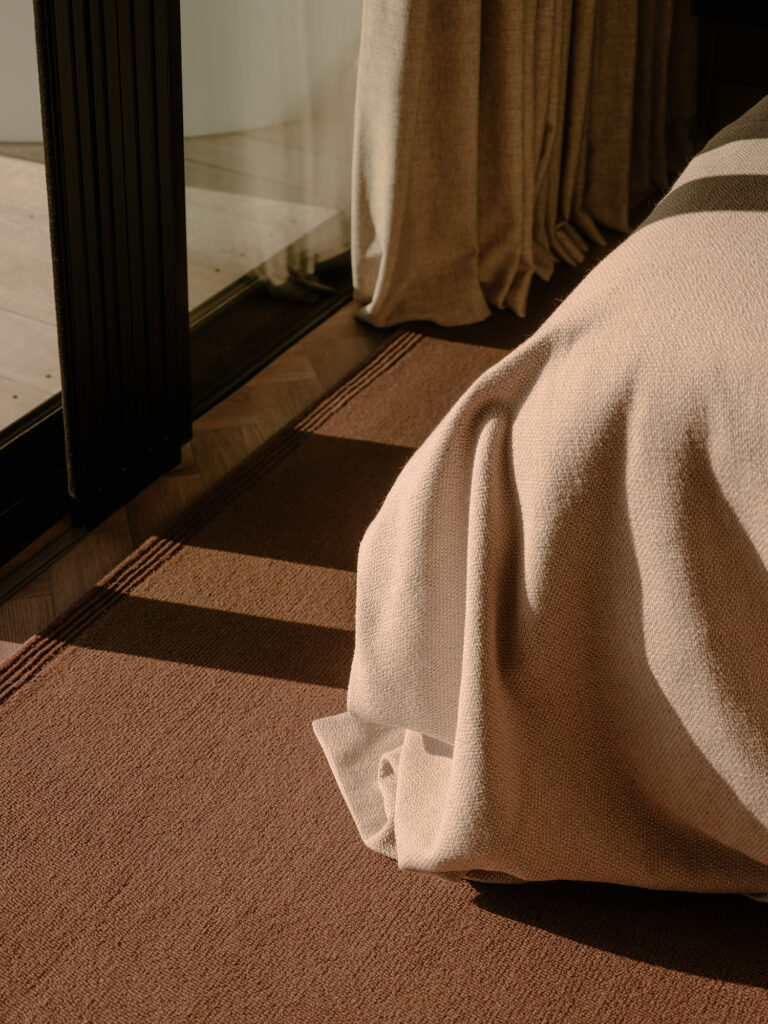 Ein Detailansicht eines Schlafzimmers mit weichen Betttextilien, warmen Farbtönen und natürlichen Stoffen vor einer Glasfront zur Terrasse